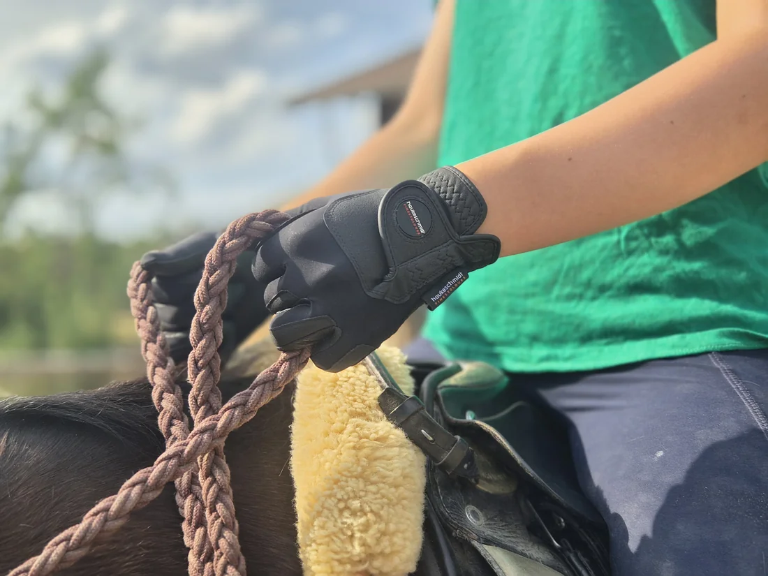 Schwarze haukeschmidt-Reithandschuhe beim Halten der Zügel auf dem Pferd